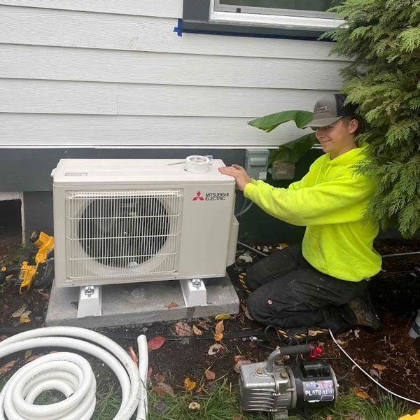 HVAC technician in yellow hoodie kneeling next to an air conditioning unit outside a home.