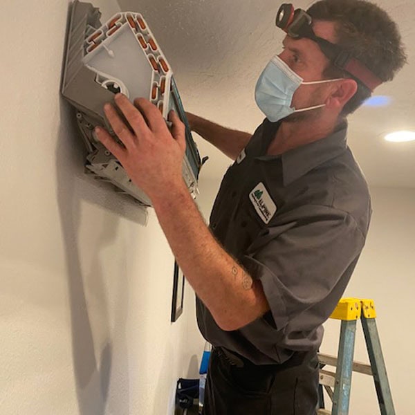 HVAC technician on a ladder repairing a ductess HVAC system on the wall of a home.