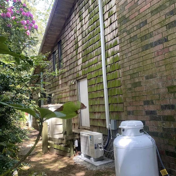 A mossy home with HVAC an system installed outside with a pink floral tree above.